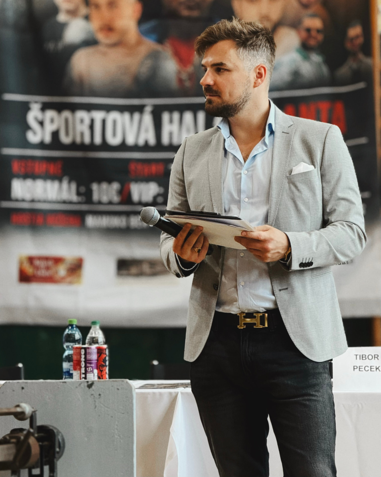 Ján Mrázik vedie tlačovú konferenciu v sivom saku s mikrofónom a poznámkami